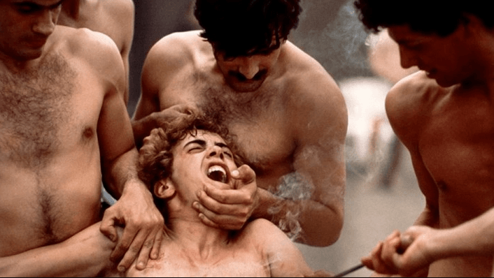 Tres chicos sin camiseta agarran a un cuarto joven de rodillas mientras le quema con un hierro hirviendo