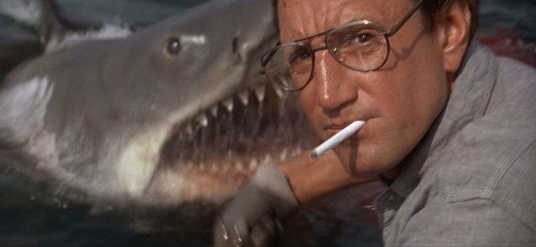 Roy Scheider en primer plano fumando un cigarrillo mirando a cámara sin darse cuenta de que el aterrador tiburón está nadando a escasos centímetros de él